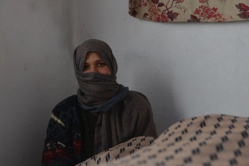 A woman wearing a grey head covering sits in the corner of a room, next to a large blanket