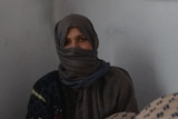 A woman wearing a grey head covering sits in the corner of a room, next to a large blanket
