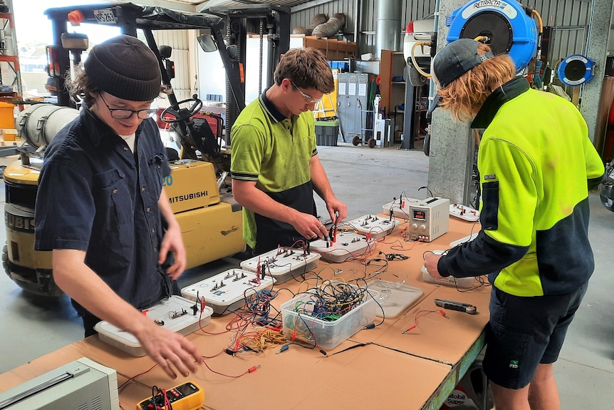 Three young men at table working on electrical circuit boards in workshop with forklift in background