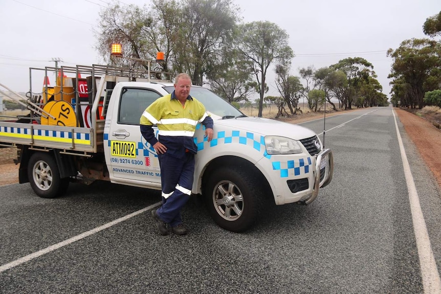 A man in high vis leans on his white ute parked across a rural stretch of road, with road closed signs in the back.