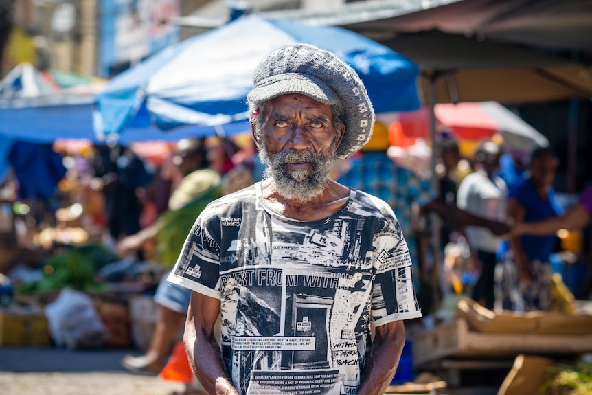 An older man with a beard and slouchy beanie stands at a marketplace 