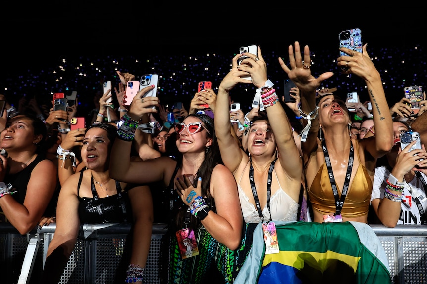Women at the front of a massive concert crowd, screaming, holding their phones, with a Brazil flag