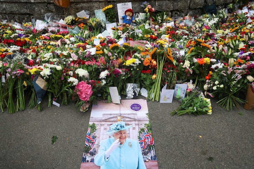 A large pile of flowers and cards on the ground, behind a poster of Queen Elizabeth II.