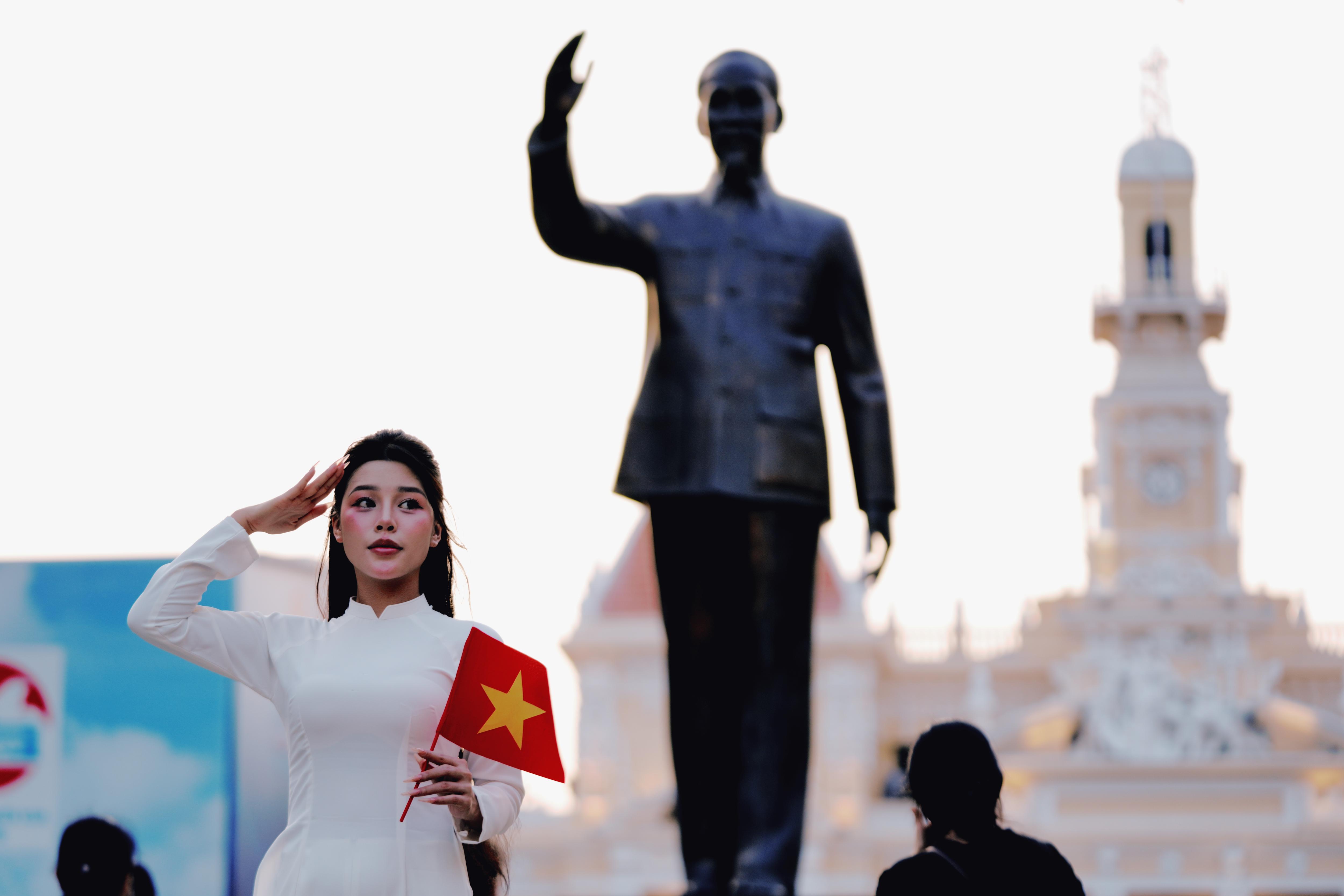 A woman in a white Ao Dai salutes while holding a Vietnamese flag near a statue of Ho Chi Minh.