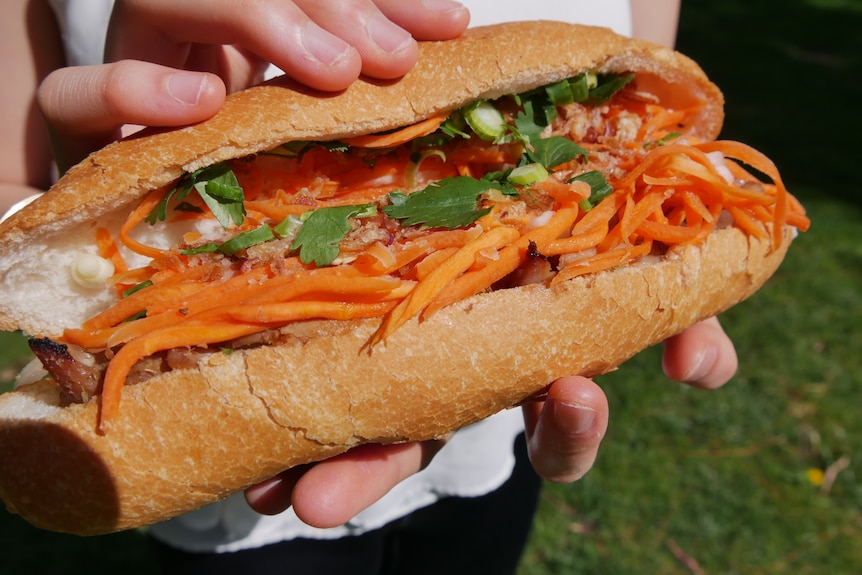 A teenage girl's hands are seen holding a bánh mì while standing in what appears to be a park.