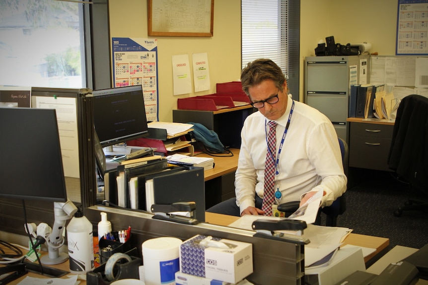 A detective sits at his desk, surrounded by folders and computers.