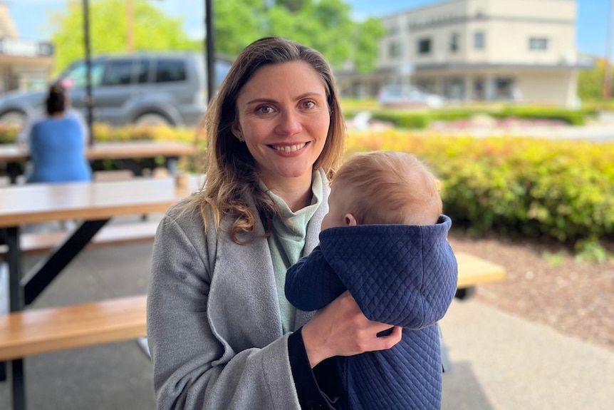 Young woman in grey coat smiles at camera holding her baby so the back of his head is visible 