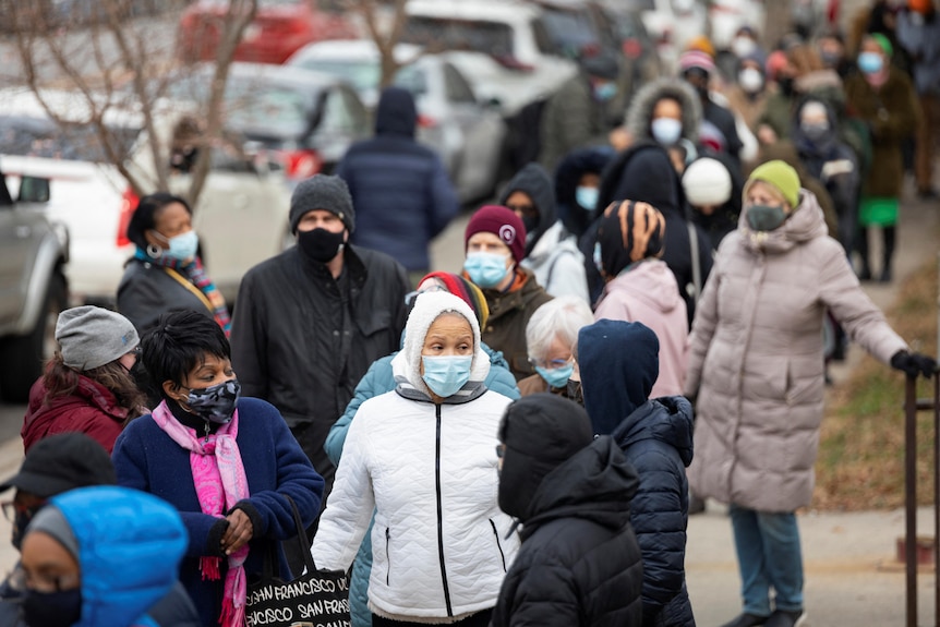A queue of people wearing face masks and winter coats and hats stand on the street, the line stretching down the block