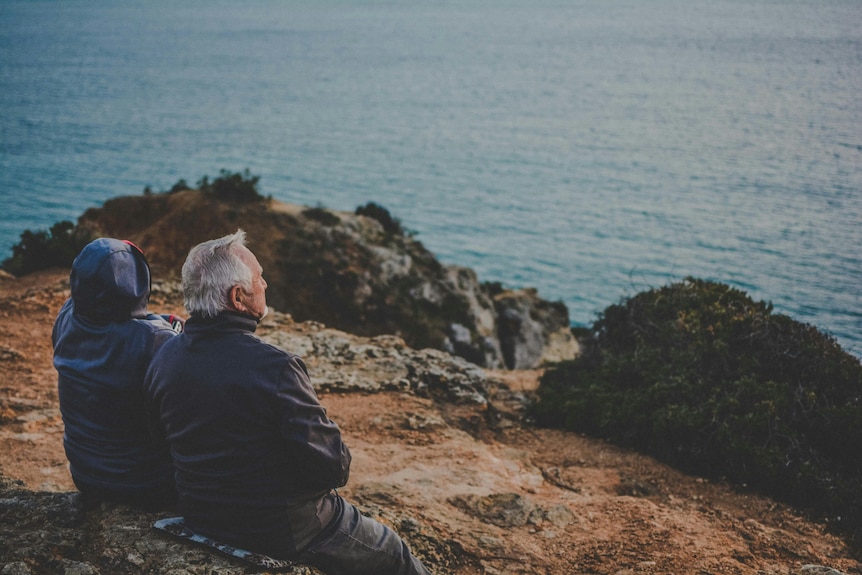 An elderly couple sits on a bench looking over a cliff with their backs to the camera.