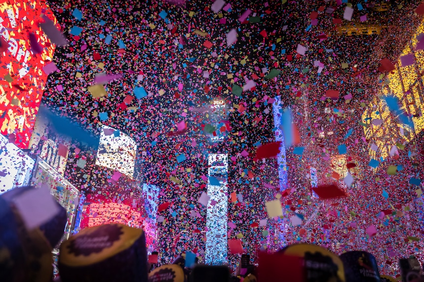 Confetti fills the sky above a busy city square that is surrounded by large buildings with electronic billboards.