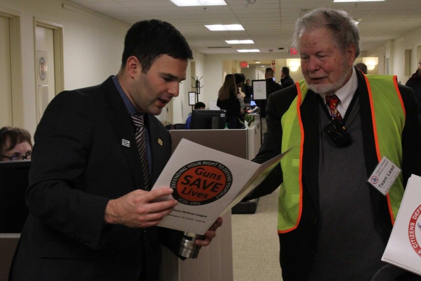 Tom Helmer holds a document with the logo Guns Save Lives on it as a man in a fluro vest looks at the document