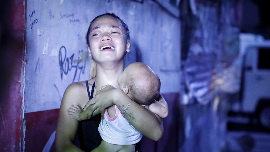 A young Asian woman in a black singlet holds a baby to her chest and cries 