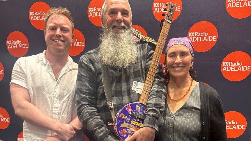 two men and one woman pose in front of black pull up banner with red dots man in the middle with long grey beard and guitar