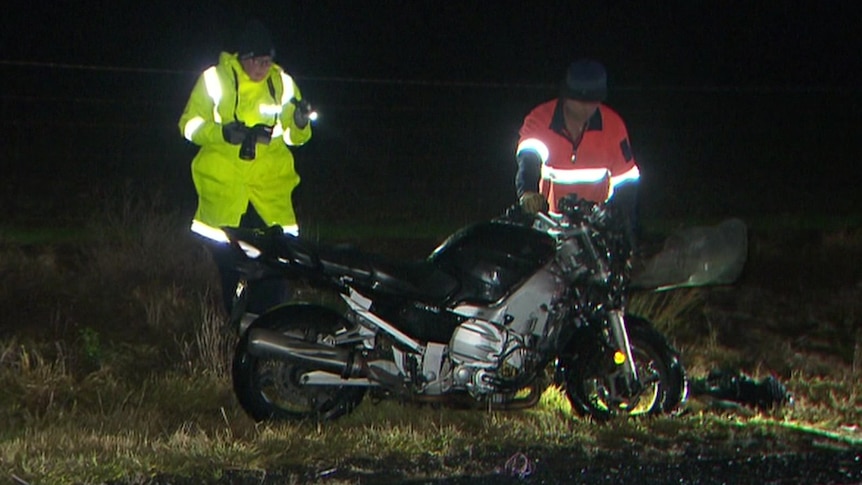 A man in yellow and a man in orange stand next to a motorcycle at night