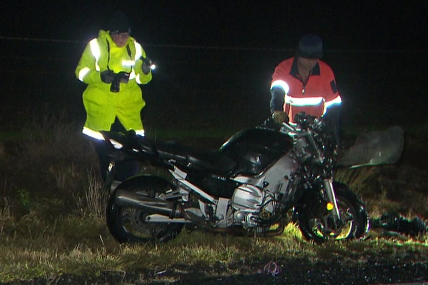 A man in yellow and a man in orange stand next to a motorcycle at night