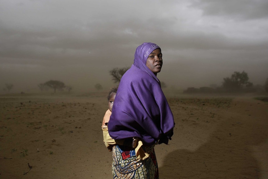 A woman with a child wrapped to her back wears a purple scarf in a dusty barren landscape.