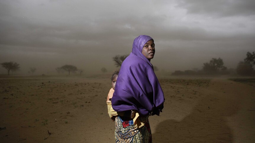 A woman with a child wrapped to her back wears a purple scarf in a dusty barren landscape.