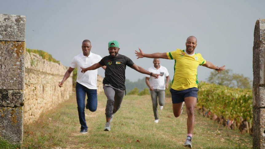 Four Zimbabwean men are sprinting towards the camera between a French vineyard and old stone wall. Three are smiling widely.