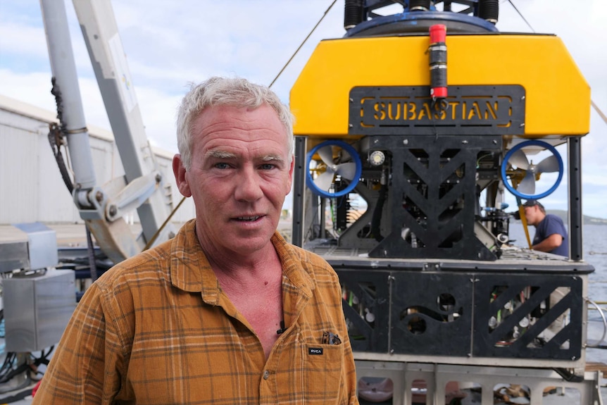 A man in an orange shirt stands on a boat in front of a submarine.