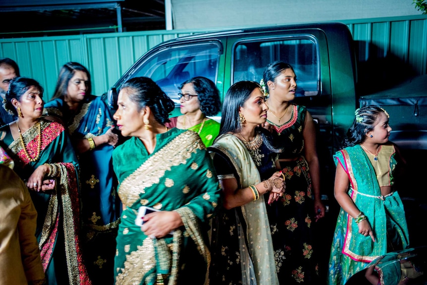 Against a green fence and with a ute in the background, several guests wait in the dress saris.