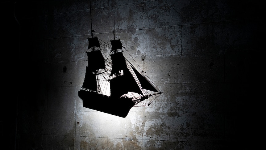 On a textured brick wall in dark room a small light creates silhouette of a wall mounted colonial vessel sculpture.