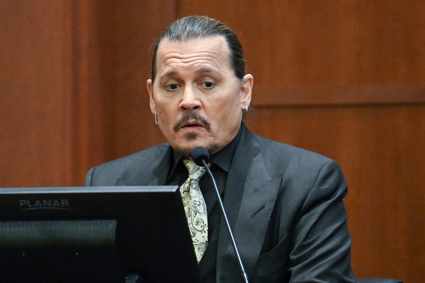 Man in a black shirt and suit with a green tie testifies in court.