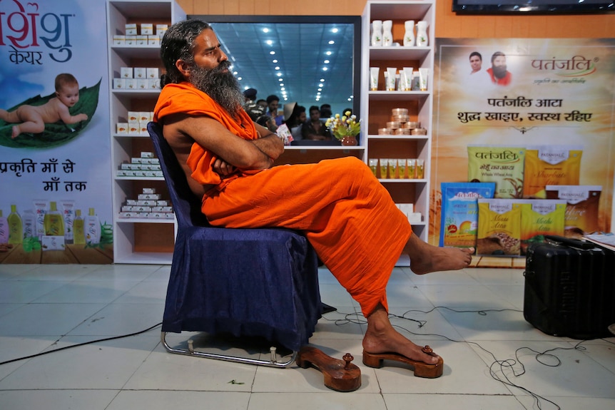 A middle-aged Indian man with a long beard sits in a chair in front of shelves of health products.
