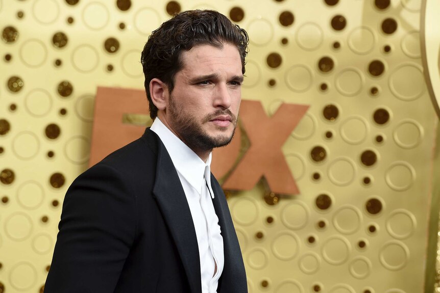 Kit Harrington looks into an unseen camera as he poses against a gold background. He wears a black suit and white shirt.