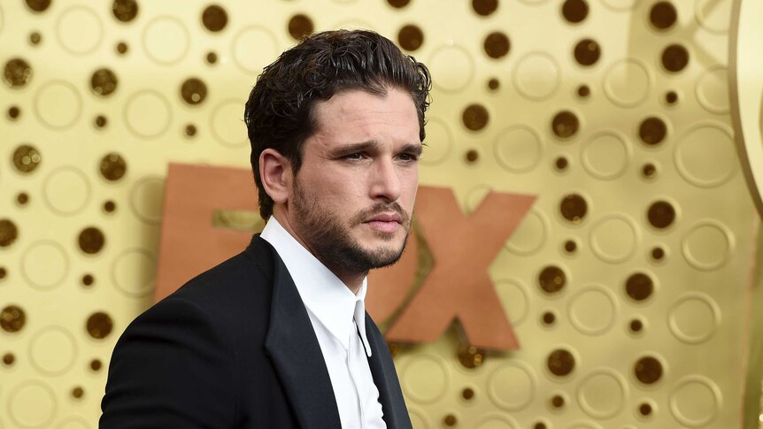 Kit Harrington looks into an unseen camera as he poses against a gold background. He wears a black suit and white shirt.