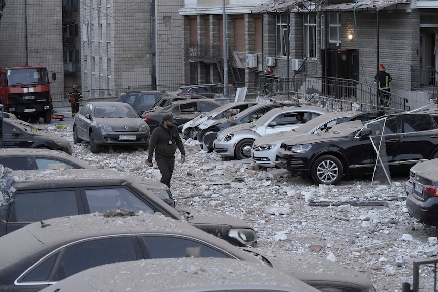 A police officer walks past damaged cars, covered with shattered glass, in front of a multi-storey apartment building.