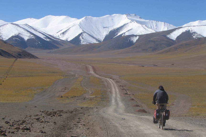 An adult on a bicycle rolls down a long unpaved road through a sparse valley towards huge mountains.