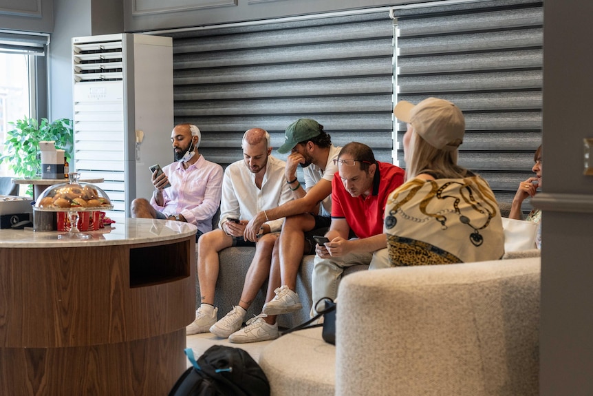 Six people sit on sofas in a waiting room, several are bald with bandages or hats