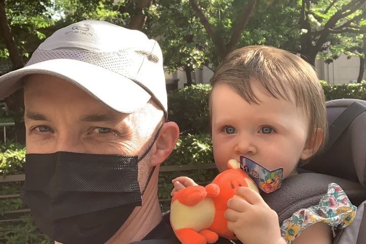 A man wearing a facial mask is taking a selfie with a baby.