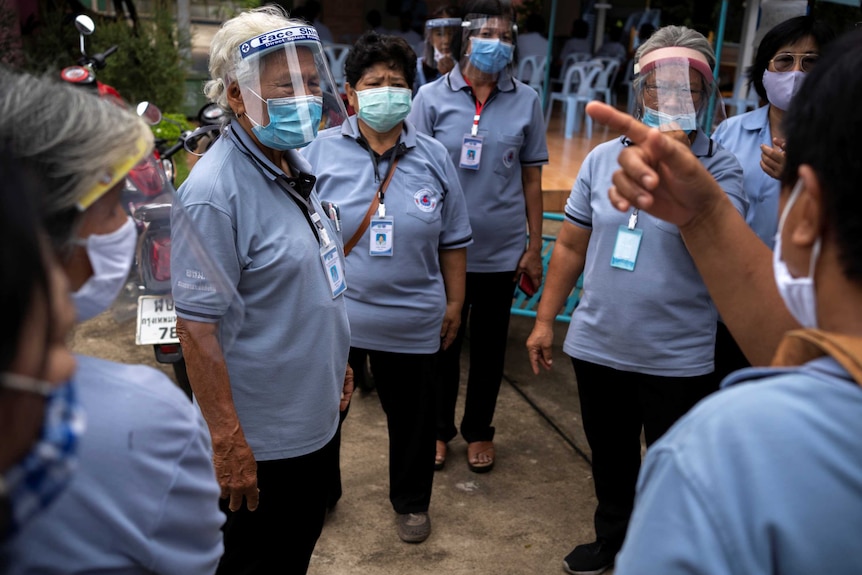 A group of older Thai women in blue polo shirts and clear facial visors