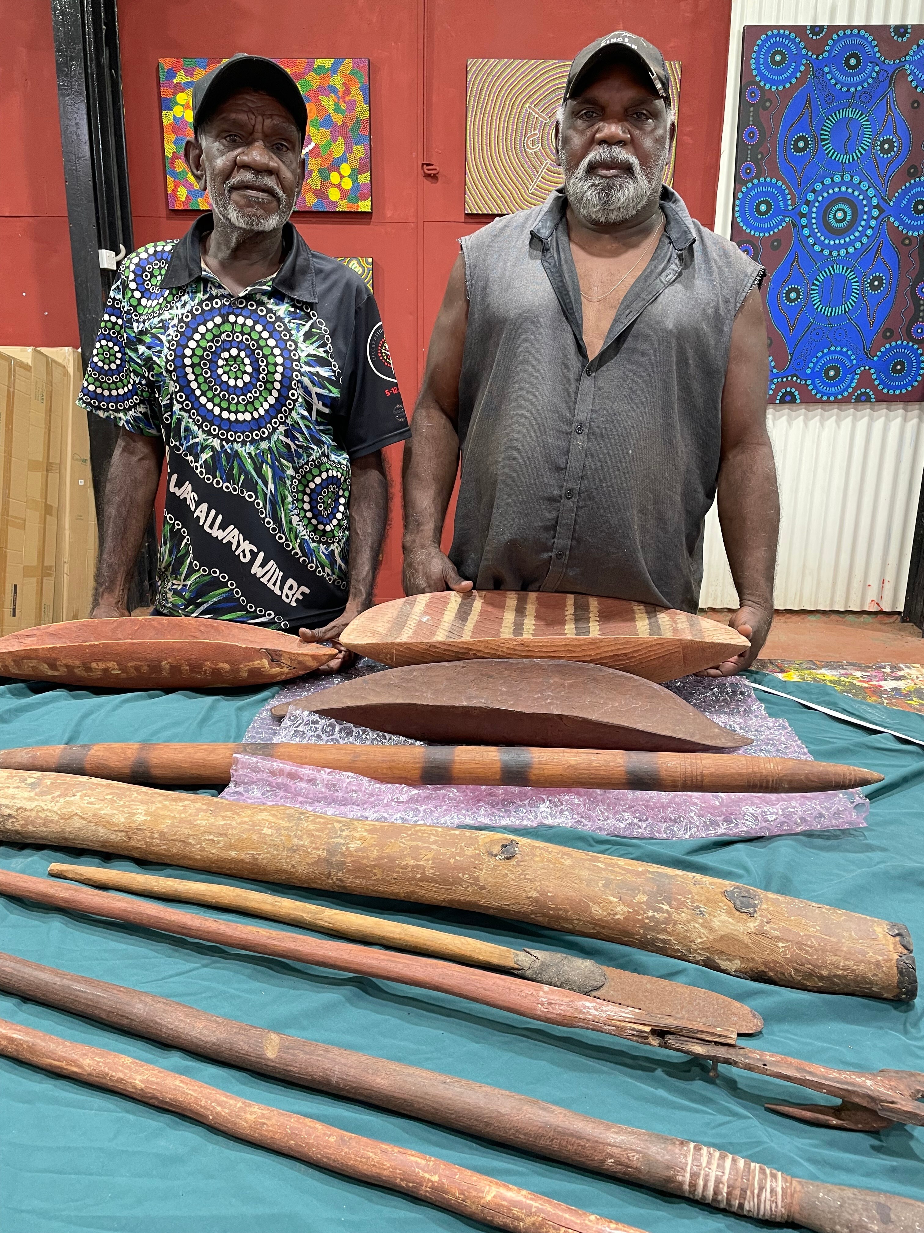 Two men stand in front of a number of Aboriginal artefacts