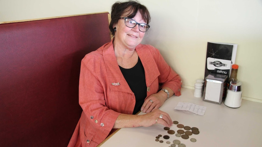 A 60-year-old woman with pre-decimal currency sits in an old-style cafe booth.
