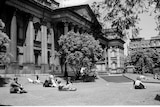 People sit in the sun on the lawn outside a public building.