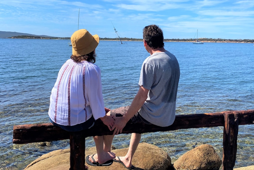 Women on left, man on right sitting on bench pole looking out to background where mast is visible out of ocean.