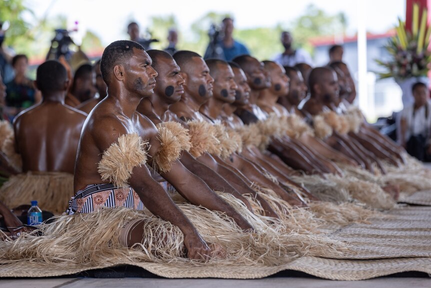 A group of Fijian men in traditional grass skirts sitting. 