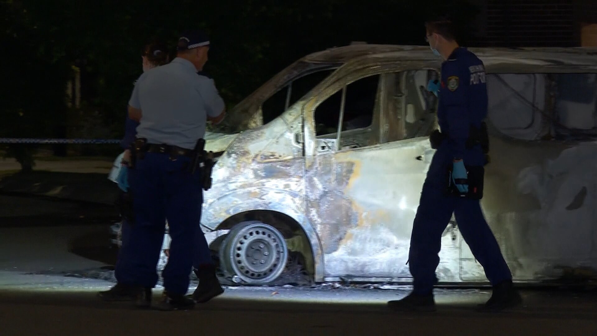 A burnt-out car with police officers looking at it with a torch, at night.