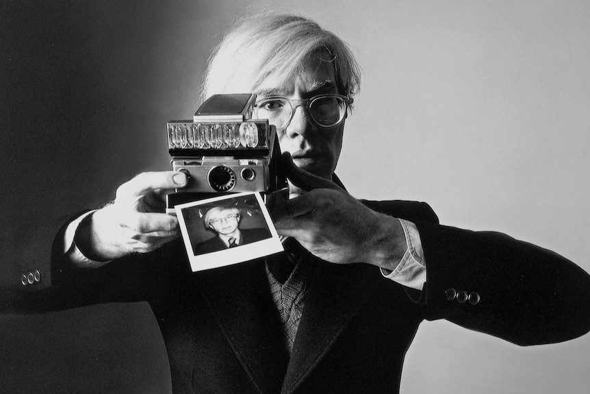 A black-and-white photograph of Andy Warhol moving a polaroid camera away from his face. The polaroid is his portrait.