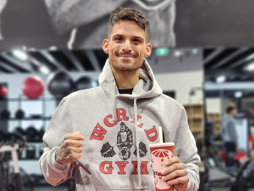 A man smiling in a boxing gym holding a drink and with clenched fist