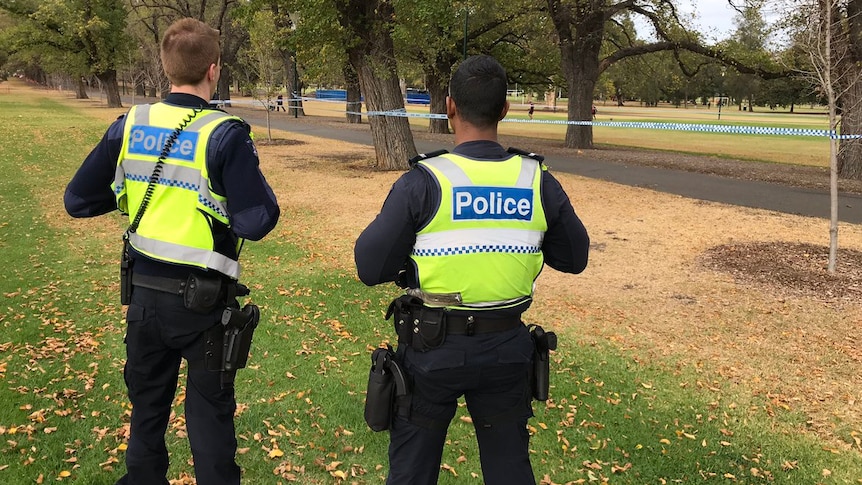 The backs of two police offices wearing fluro vests in Fawkner Park at the crime scene.
