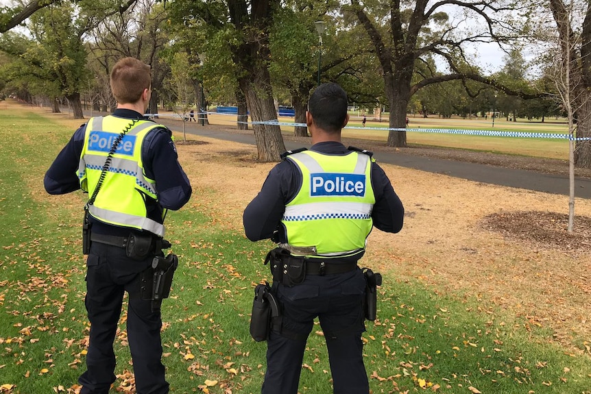 The backs of two police offices wearing fluro vests in Fawkner Park at the crime scene.