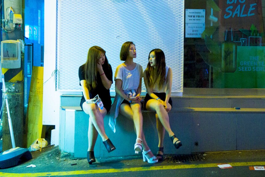 Three woman sit chatting on a street corner outside a Korean nightclub