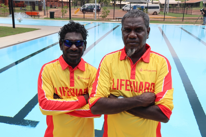 Two men in bright red and yellow lifeguard uniforms stand by a blue wimming pool and look at the camera, arms folded.