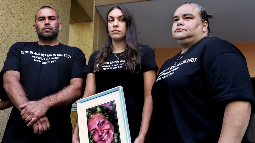 Three family members, one man and two women stand wearing black t-shirts holding a photo of Tanya Day outside a grey building