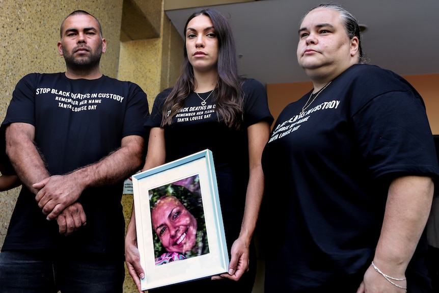 Three family members, one man and two women stand wearing black t-shirts holding a photo of Tanya Day outside a grey building