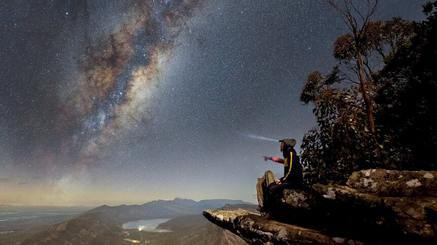 Man on rock shelf looks over valley as stars shine above