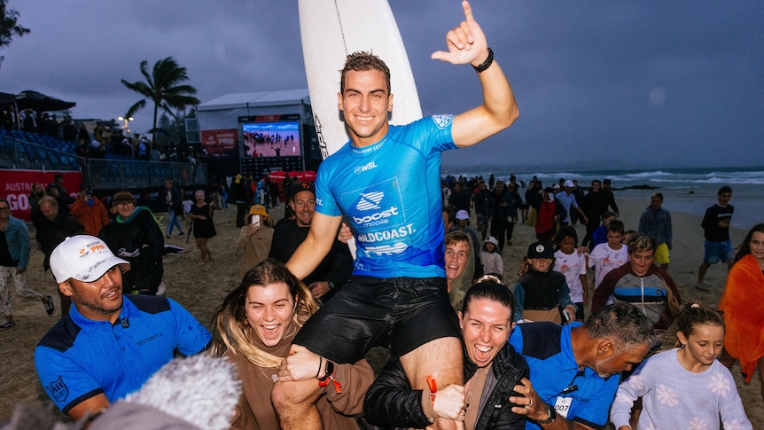 A man is grinning at the camera, held on the shoulders of two cheering women. A white surfboard and crowd is behind him.